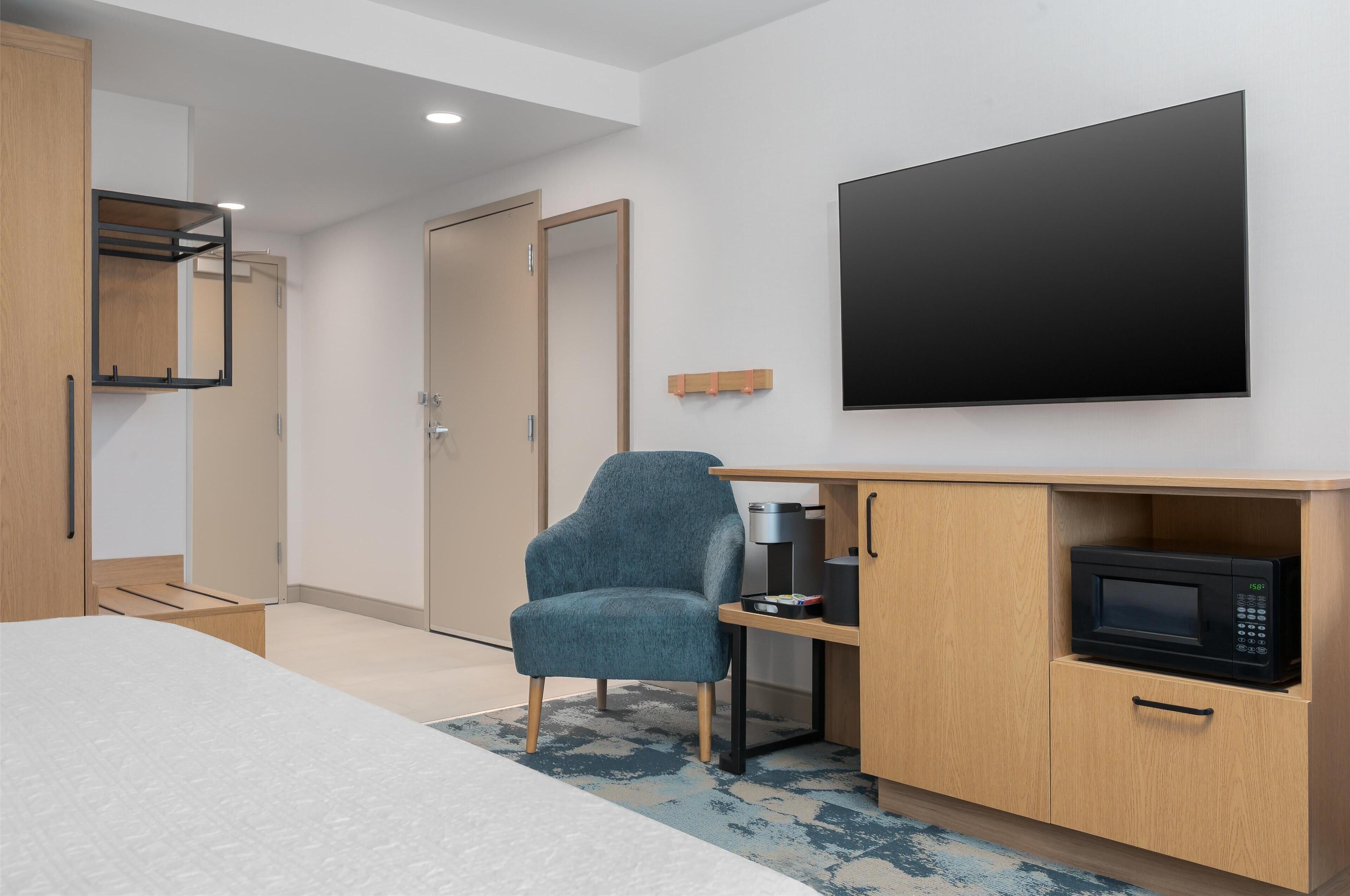A modern hotel room with a bed, a desk with a microwave, a chair, and a TV mounted on the wall.