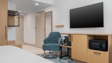 A modern hotel room with a bed, a desk with a microwave, a chair, and a TV mounted on the wall.