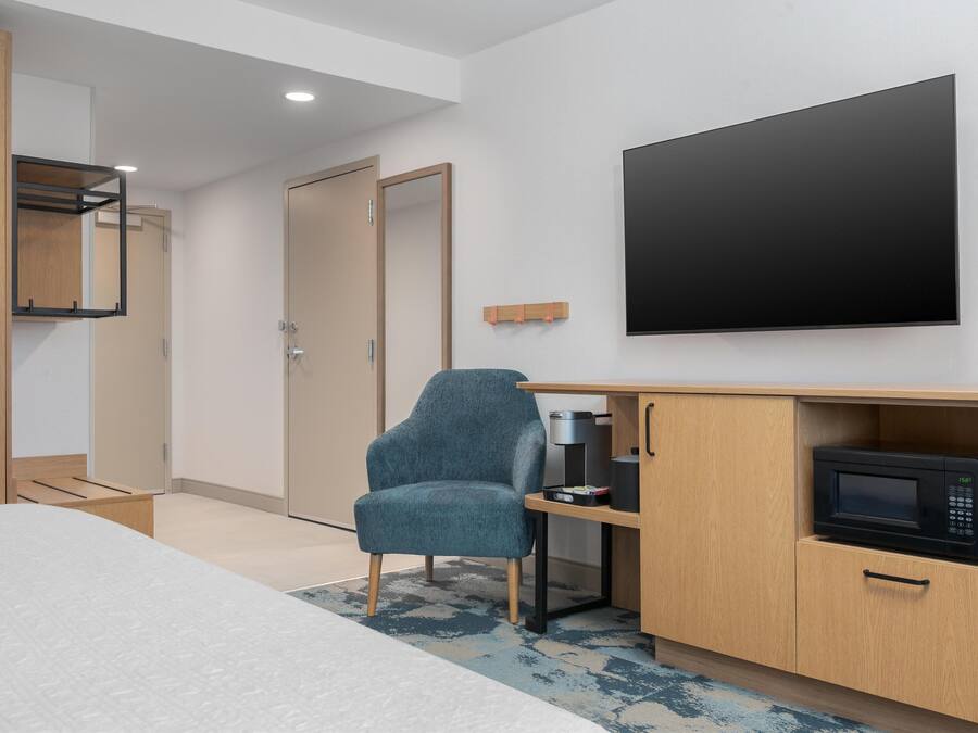A modern hotel room with a bed, a desk with a microwave, a chair, and a TV mounted on the wall.