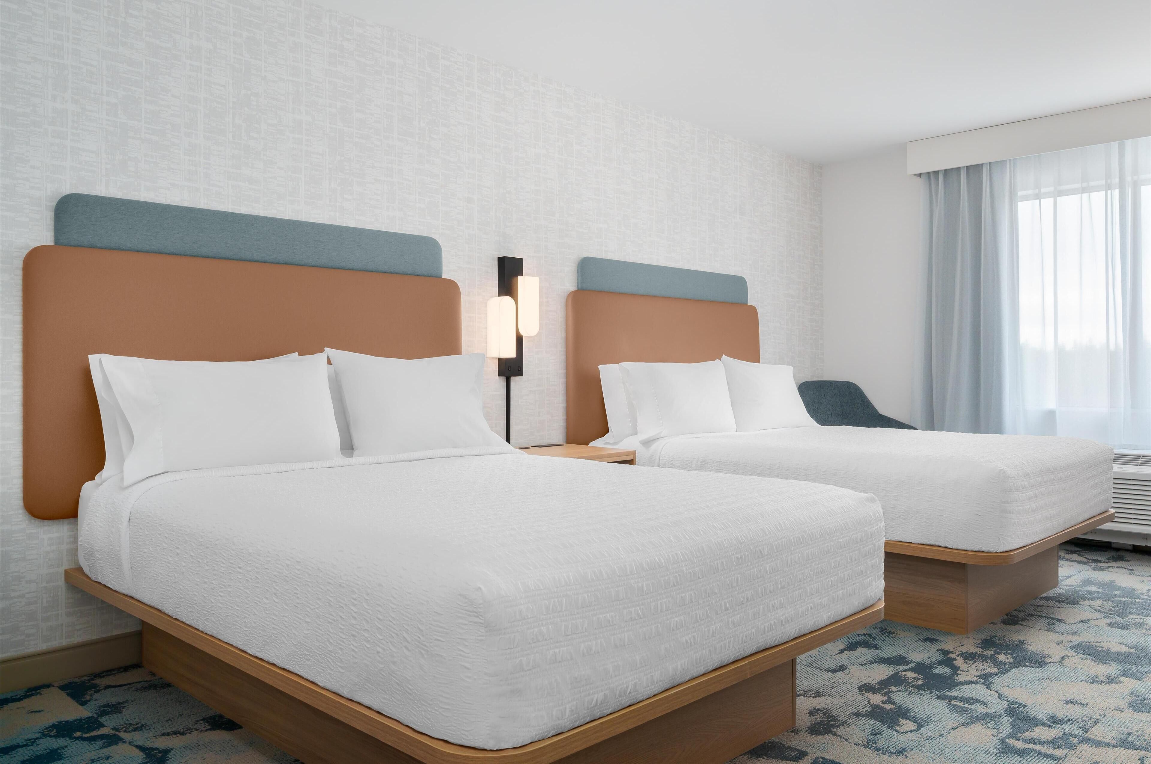 A hotel room with two beds, a nightstand, a lamp, and a window with curtains.