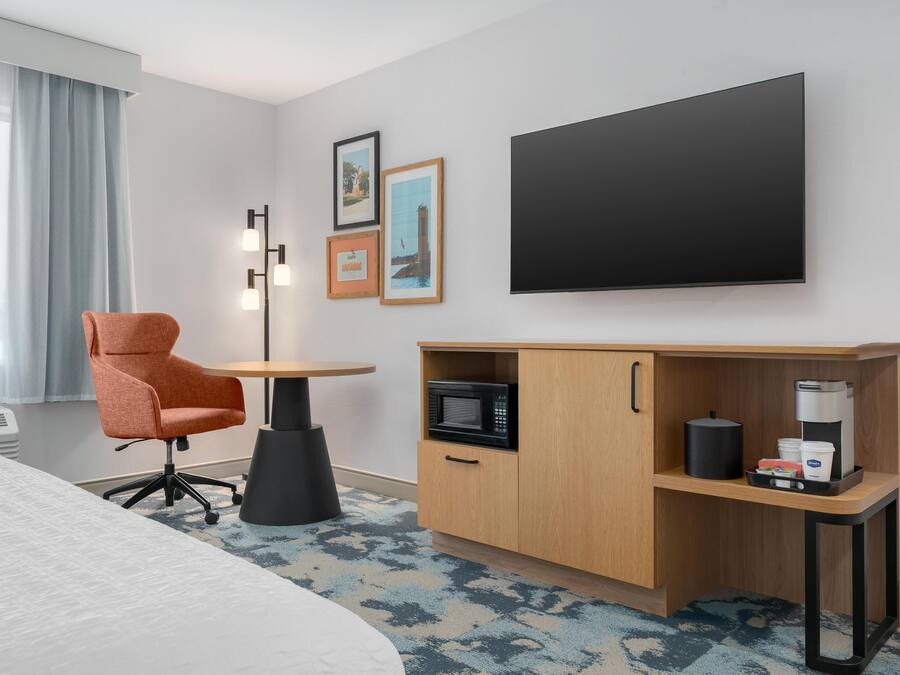 A hotel room with a bed, a desk with a microwave, a coffee maker, and a chair.
