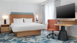 A hotel room with a bed, a desk with a chair, a TV, and a nightstand with a lamp.