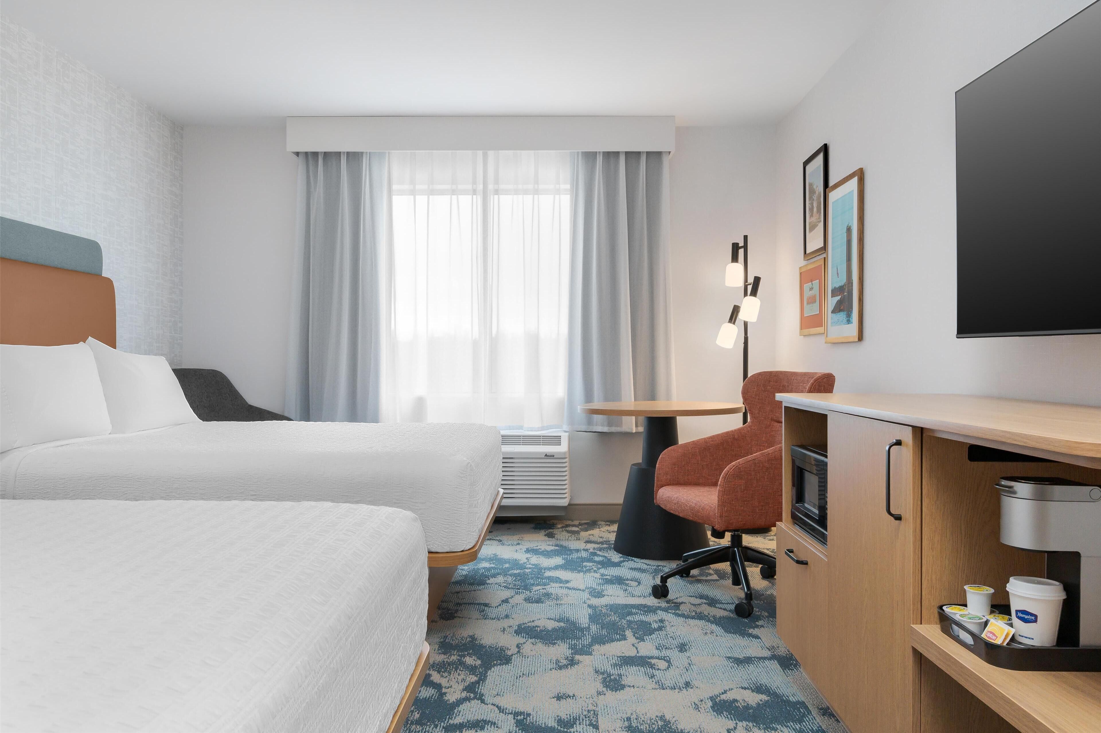 Habitación de hotel con dos camas, un escritorio, una silla, un televisor de pantalla plana, un fabricante de café y una ventana con cortinas.