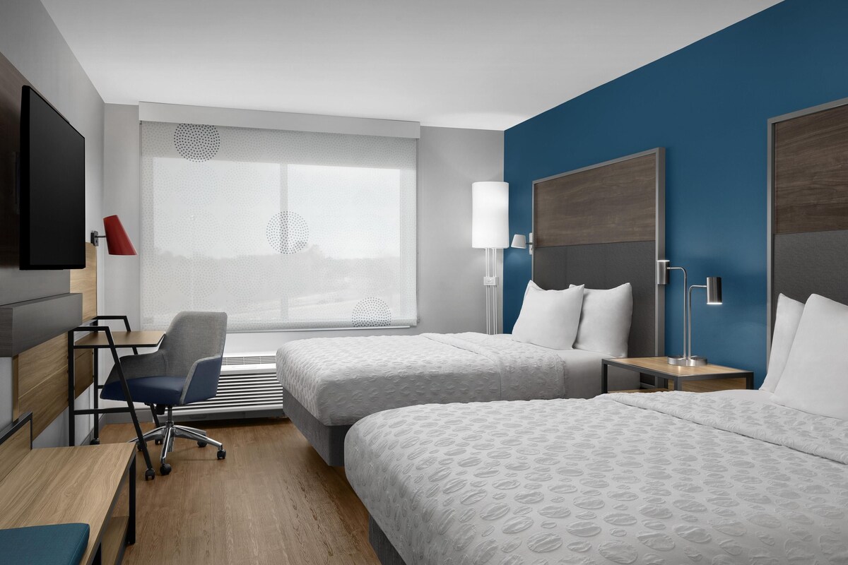 A hotel room with two beds, a desk, a chair, a TV, and a window with blinds.