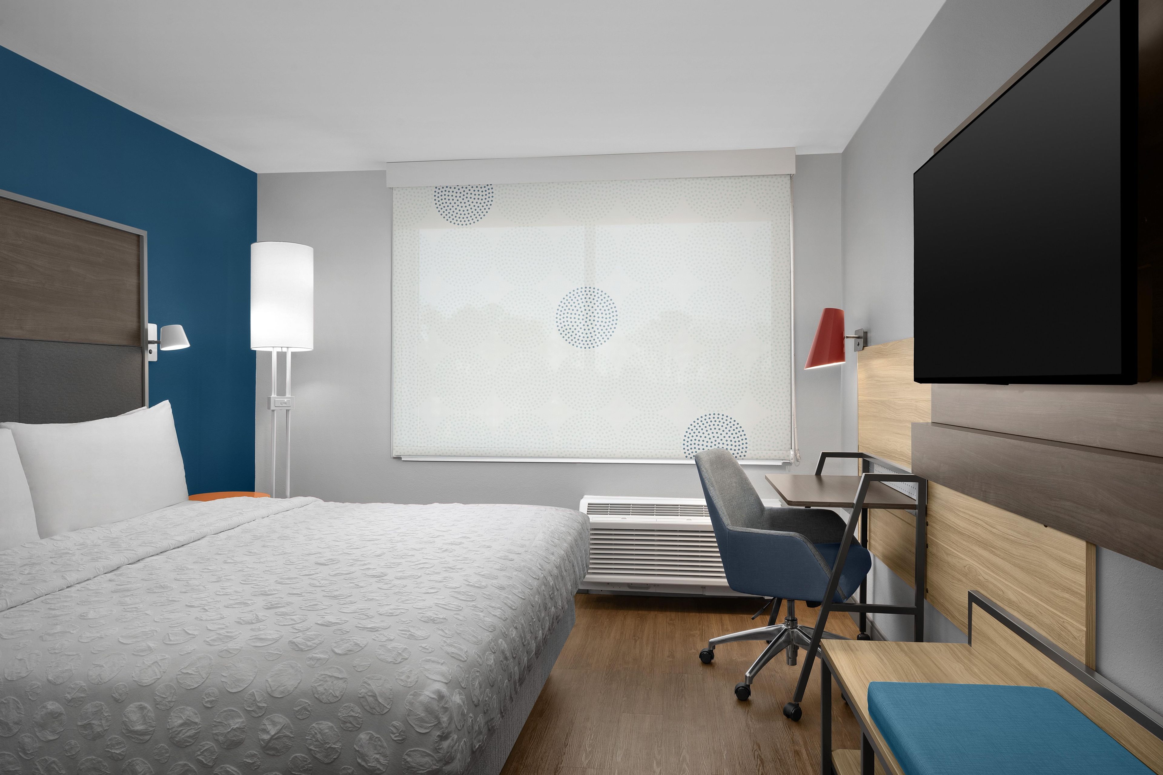 A hotel room with a bed, a desk, a chair, a TV, and a window with blinds.