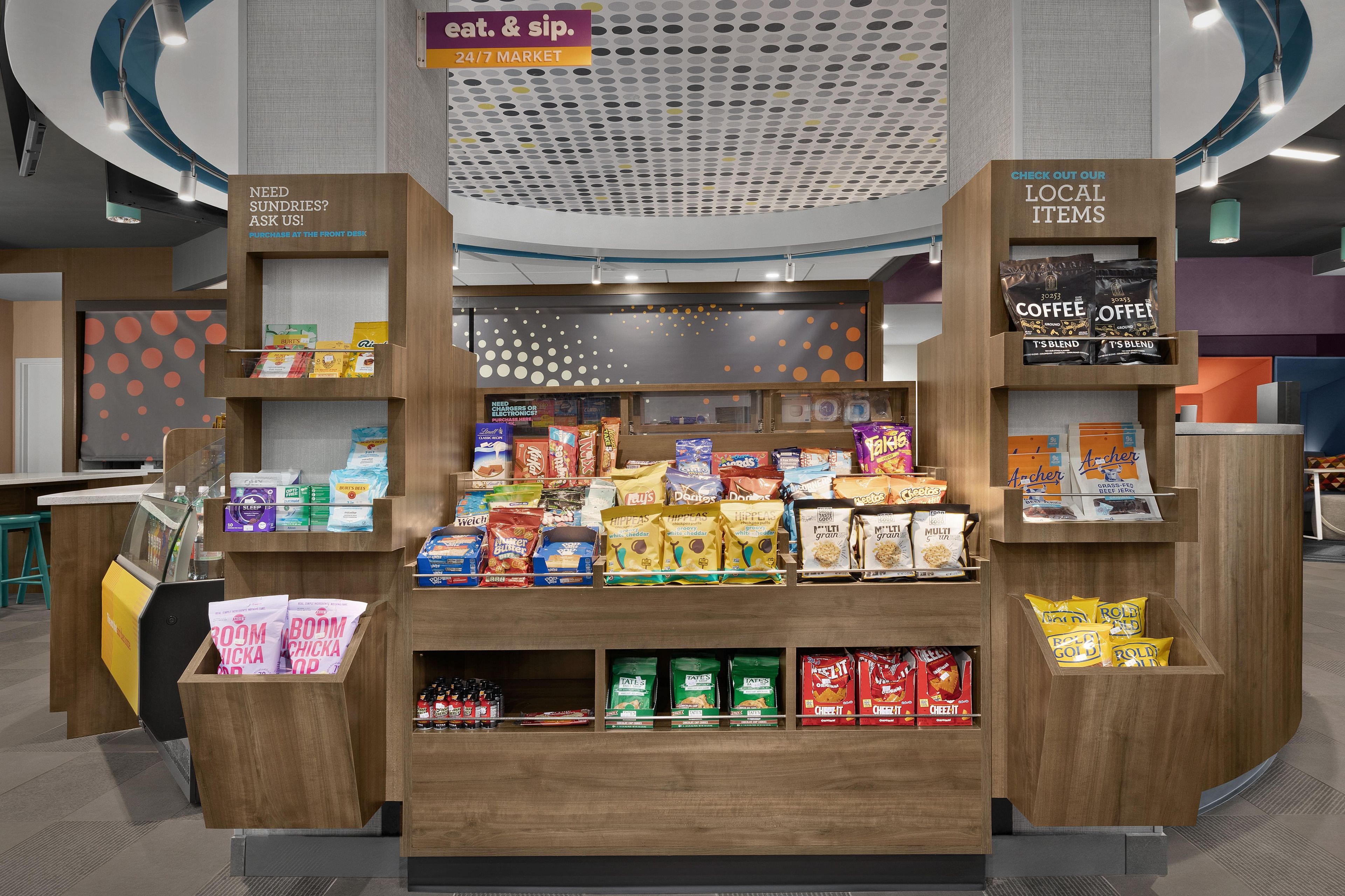 A snack display with various packaged goods, a coffee station, and a sign indicating a 24/7 market.