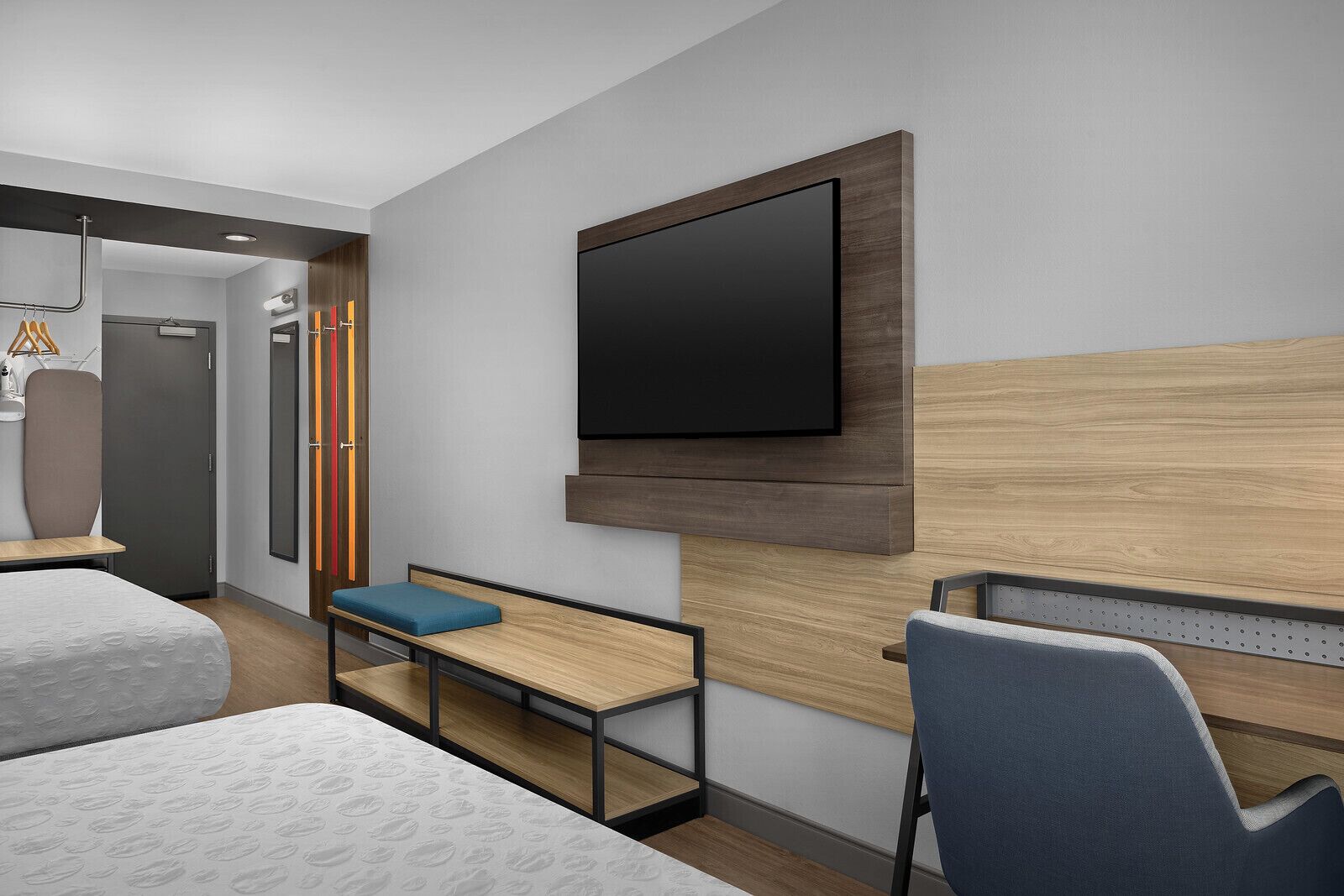 Une chambre d’hôtel moderne dotée d’un grand téléviseur à écran plat fixé au mur, d’une tête de lit en bois, d’un bureau avec une chaise et d’un lit agrémenté d’un coussin bleu.