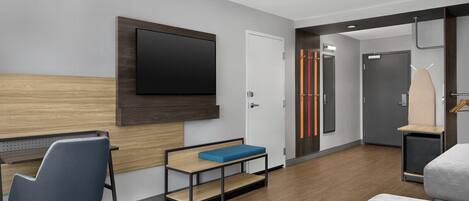 A modern hotel room with a wooden headboard, a flat-screen TV, a desk with a chair, a bench, a bed, and a wardrobe.