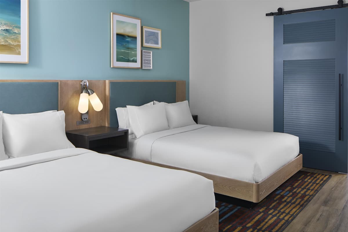 Ein Hotelzimmer mit zwei Einzelbetten, einem Nachttisch mit Lampe, einer blauen Tür und gerahmten Bildern an der Wand.