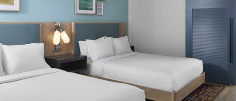 A hotel room with two single beds, a nightstand with a lamp, a blue door, and framed artwork on the wall.