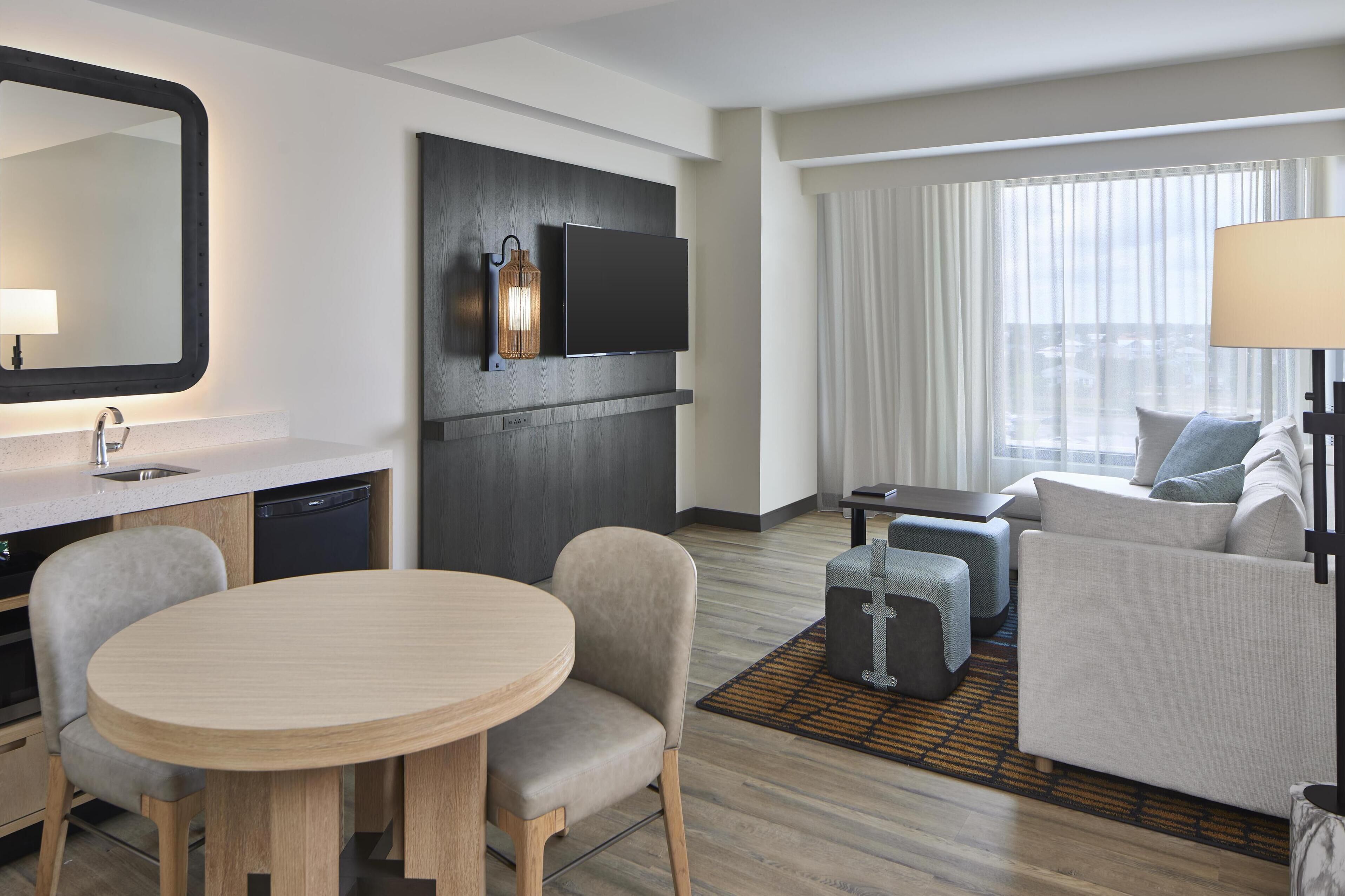 A modern hotel room with a dining area, a sofa set, a flat-screen TV, and a large window with curtains.