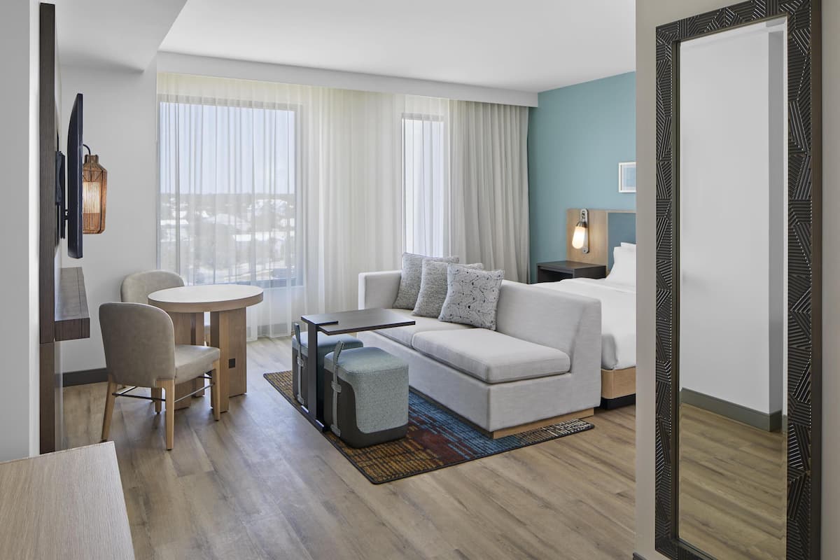 Ein modernes Hotelzimmer mit einer Couch, einem runden Tisch und einem Sessel. Es gibt ein großes Fenster mit Vorhängen, eine Lampe auf dem Nachttisch und einen Spiegel mit geometrischem Rahmen.