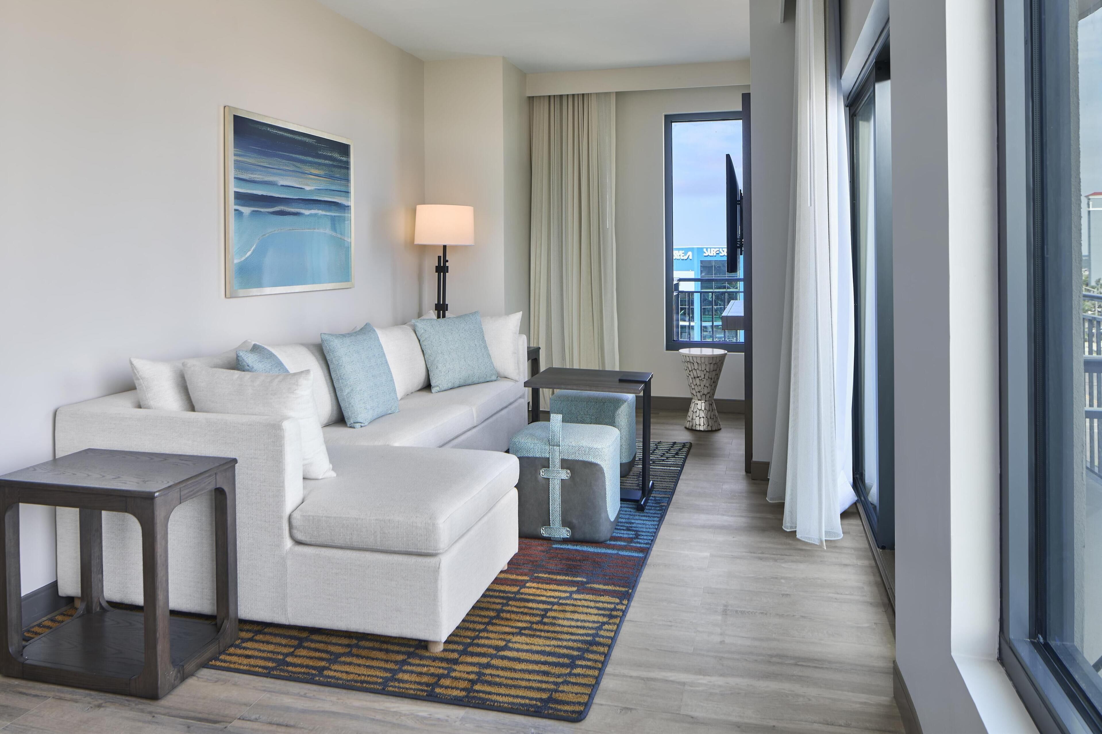 A modern hotel room with a sofa, ottoman, side table, and a view of the city through large windows.