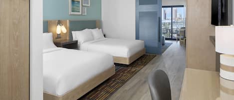 A hotel room with two beds, a desk, a chair, a TV, and a view of the city.