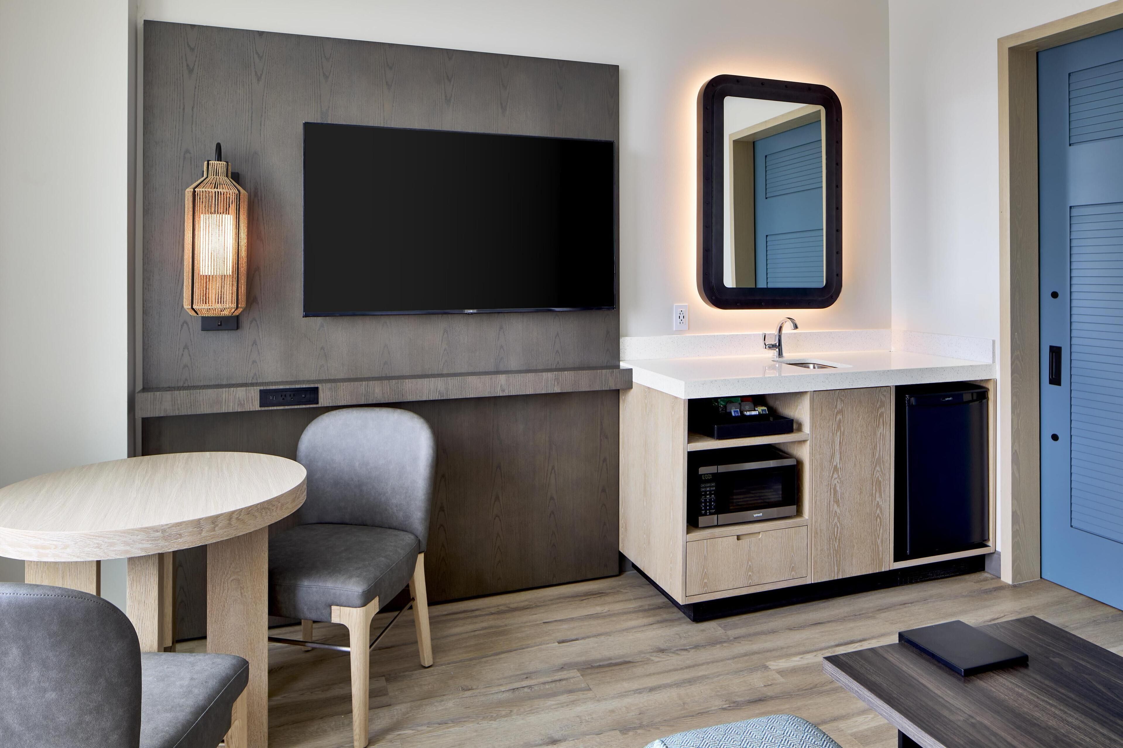A modern hotel room with a flat-screen TV, a wooden cabinet with a sink, a dining table with chairs, and a small side table.