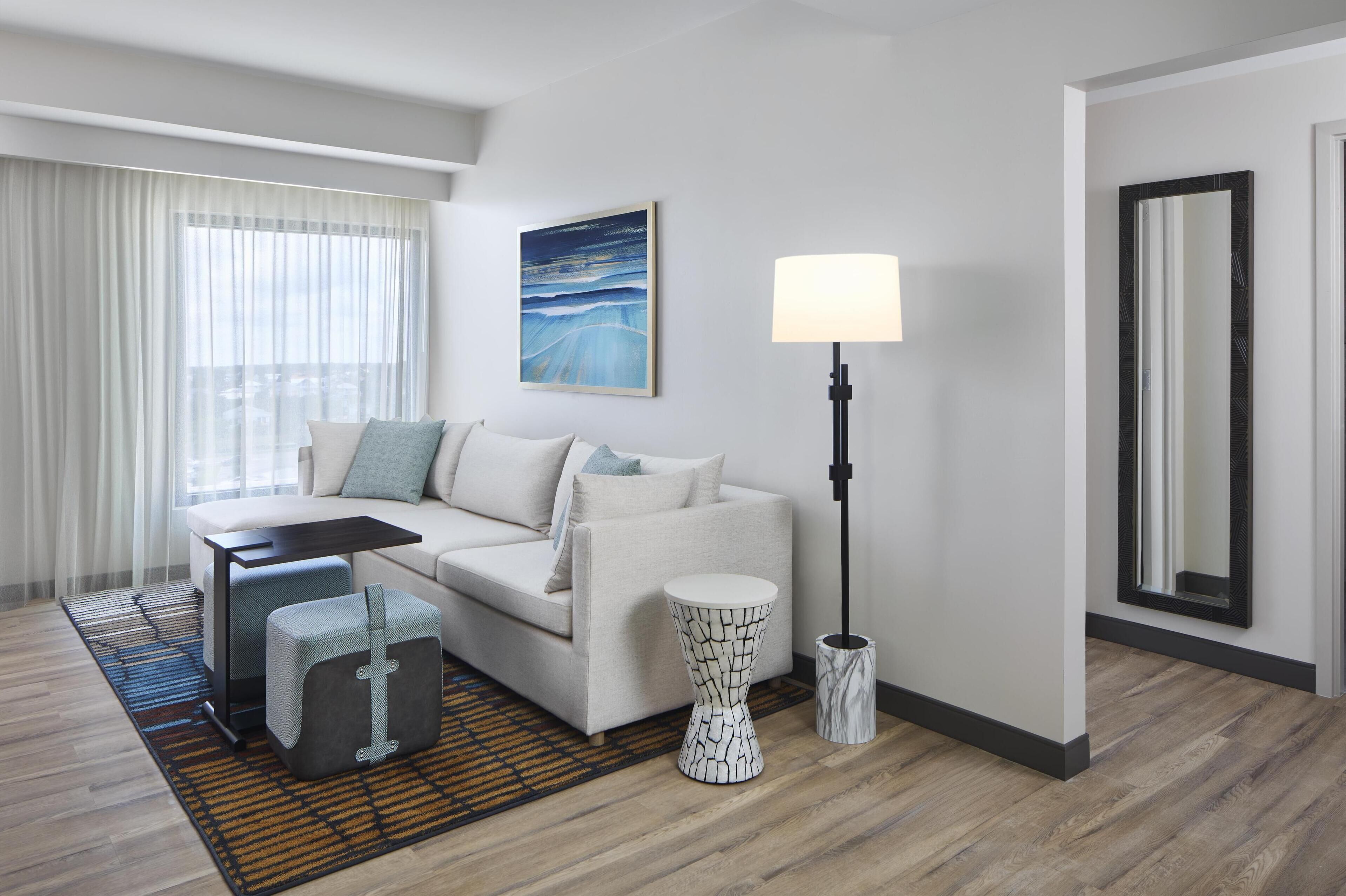 A modern hotel room with a white sofa, a small side table, a floor lamp, and a large window with sheer curtains.
