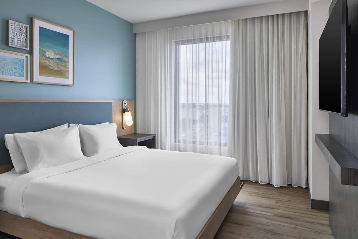 Ein Hotelzimmer mit einem Bett, einem Nachttisch mit Lampe, einem Fernseher und einem großen Fenster mit Vorhängen.