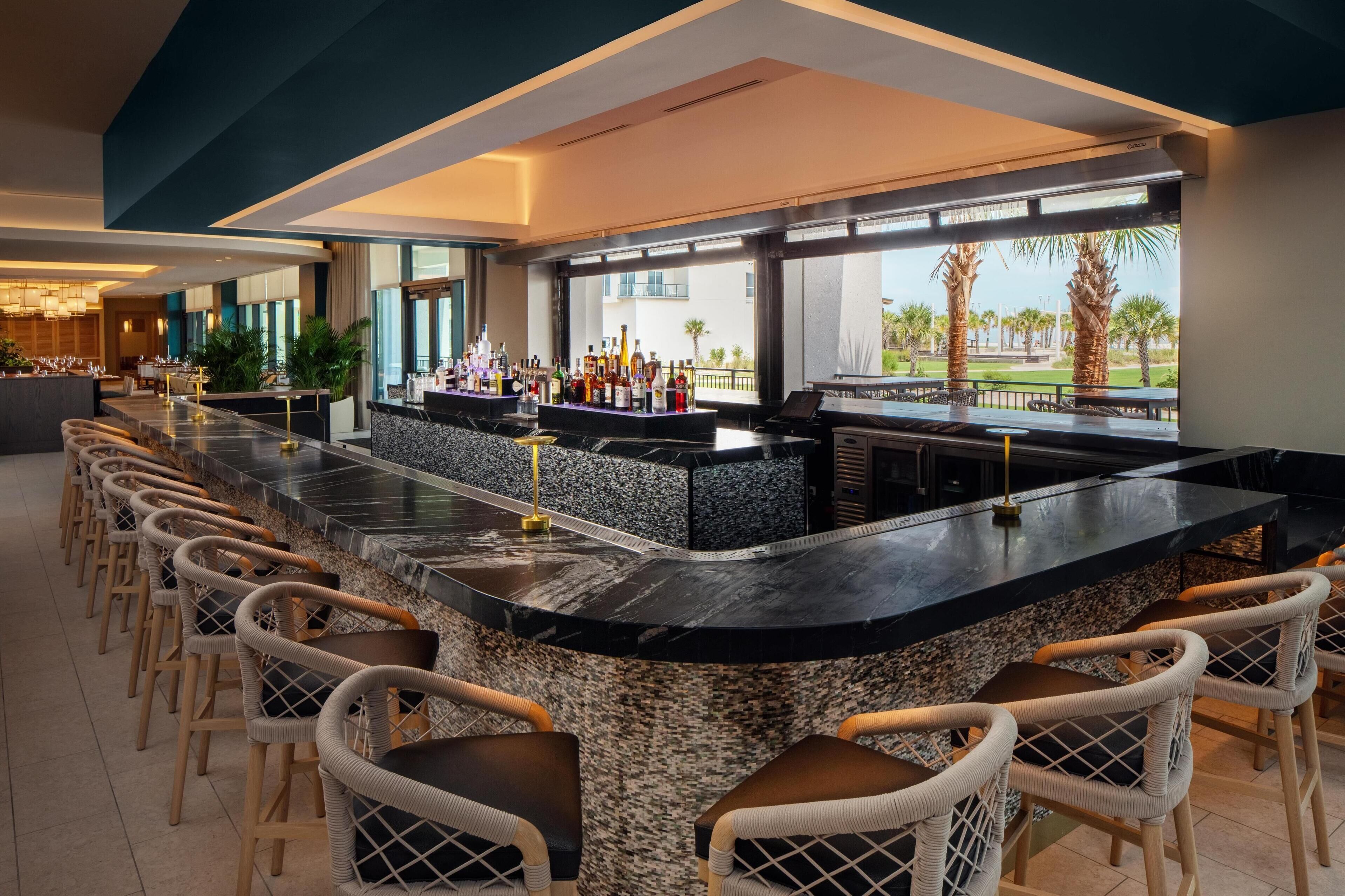 A bar area with a long counter, multiple seating options, and a view of palm trees and buildings.