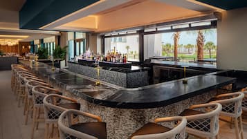 A bar area with a long counter, multiple seating options, and a view of palm trees and buildings.
