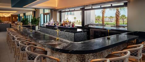 A bar area with a long counter, multiple seating options, and a view of palm trees and buildings.