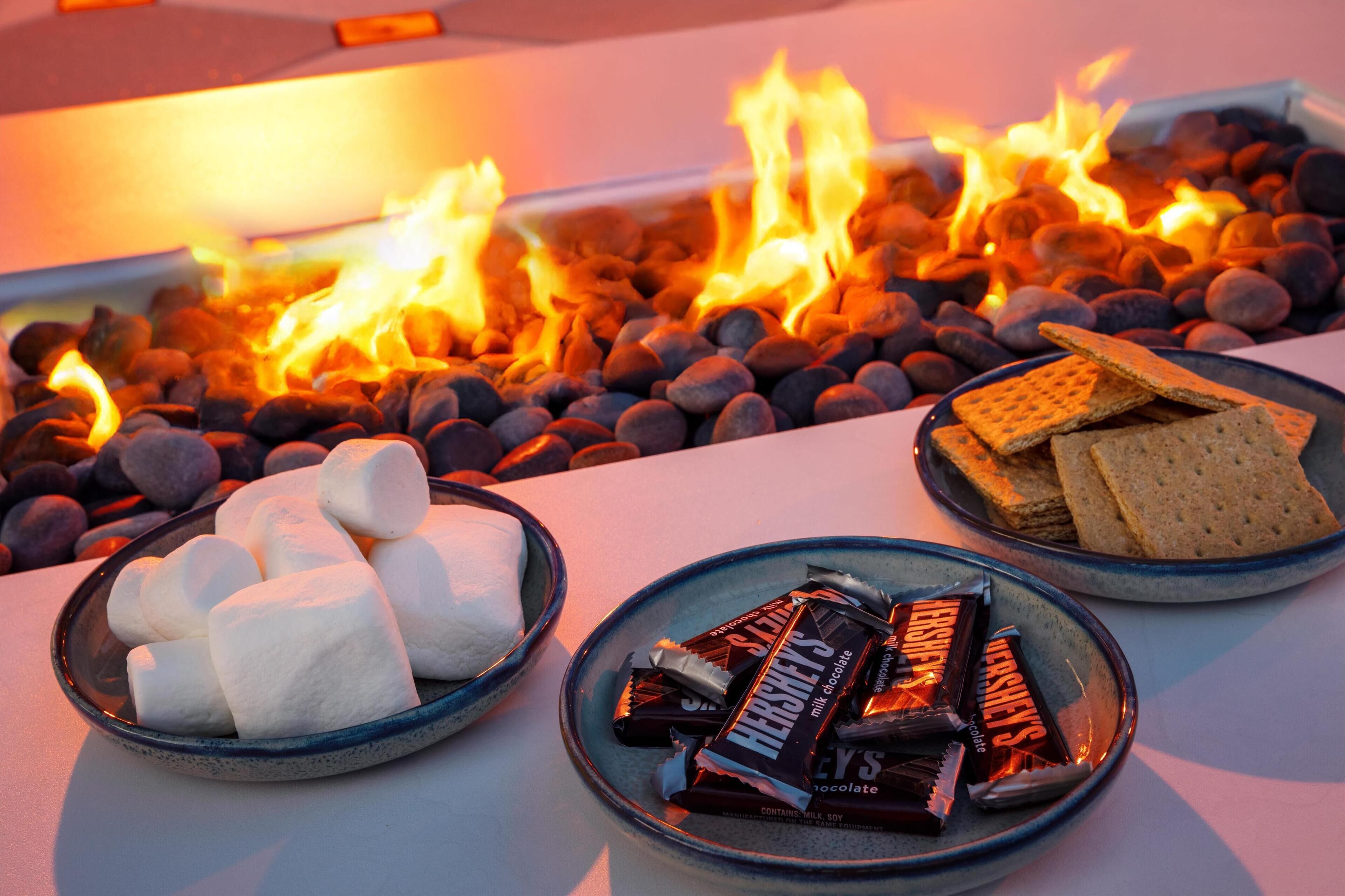 Una mesa con malvaviscos, barras de chocolate y galletas alrededor de una fogata.