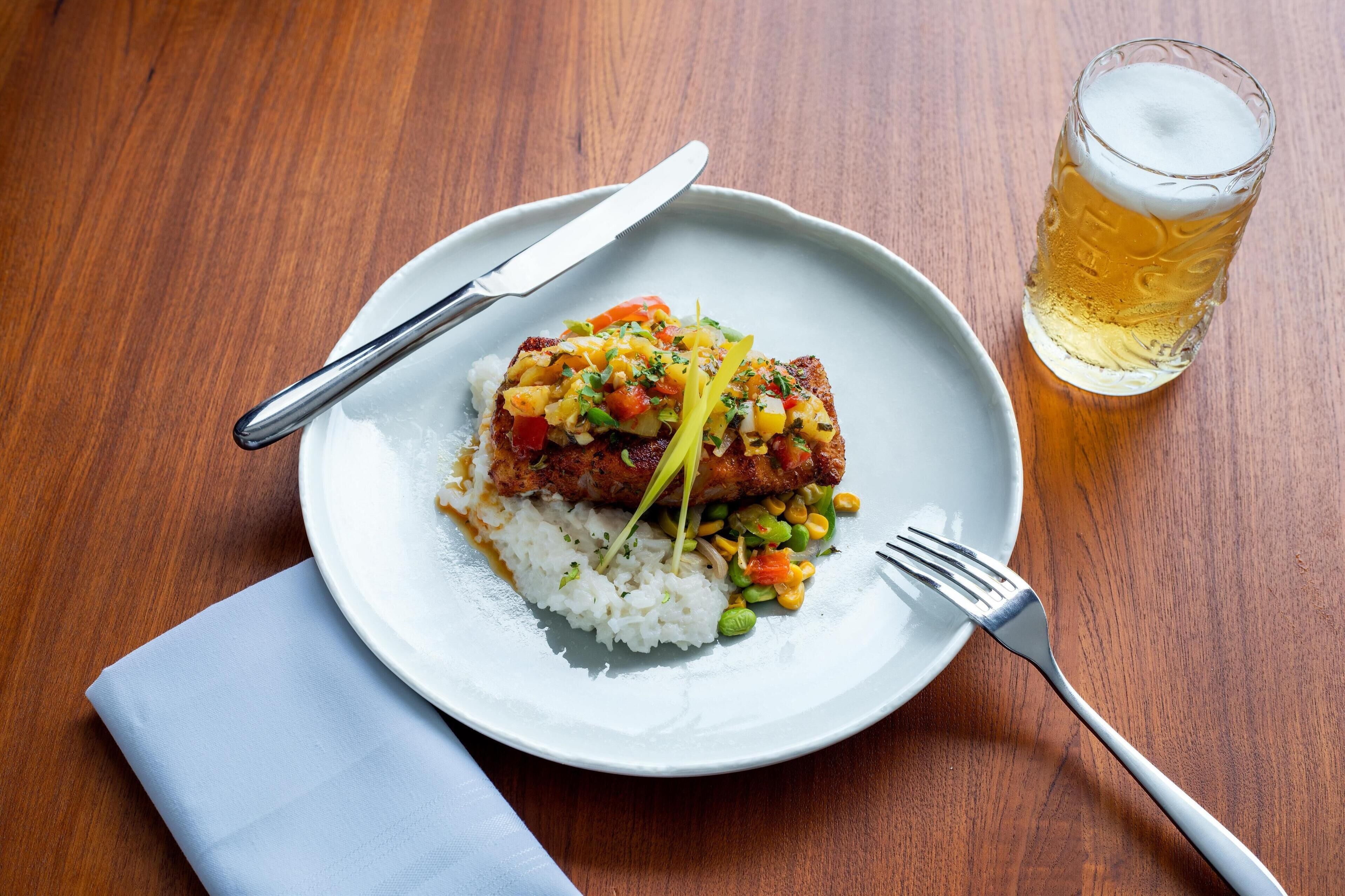 A plate of food with rice, vegetables, and a piece of meat, accompanied by a glass of beer and cutlery on a wooden table.