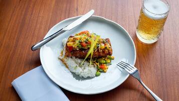 Un plato de comida con arroz, verduras y un trozo de carne, acompañado de una cerveza y cubiertos sobre una mesa de madera.