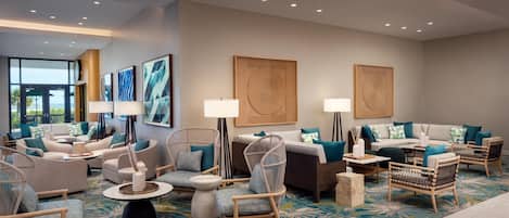 A modern hotel lobby with a seating area featuring sofas, chairs, and a coffee table. There are framed artworks on the walls and a large window providing a view outside.
