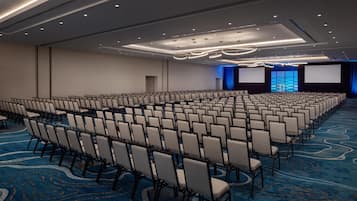 Un amplio salón de conferencias con filas de sillas, una alfombra azul y un escenario con un telón de fondo azul.