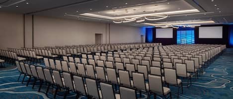 Un amplio salón de conferencias con filas de sillas, una alfombra azul y un escenario con un telón de fondo azul.