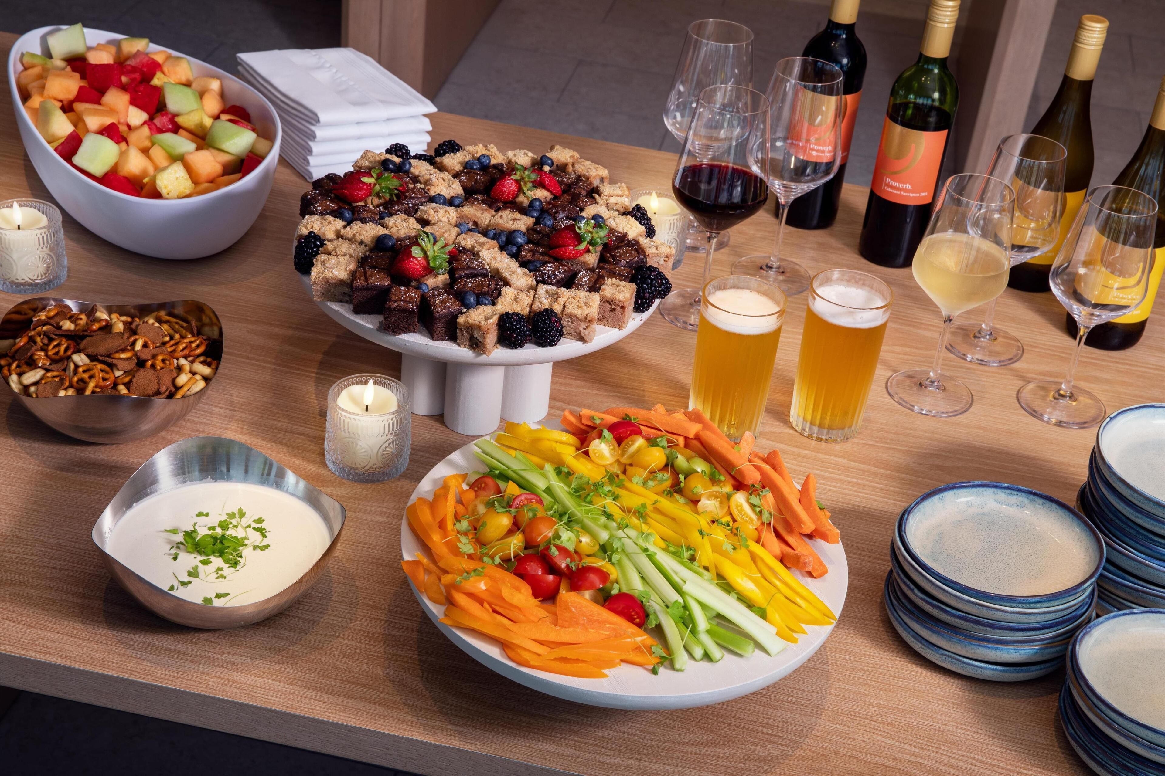 A table with various food items, including a fruit salad, a dessert tray, and a plate of vegetables. There are also several glasses of wine and beer.