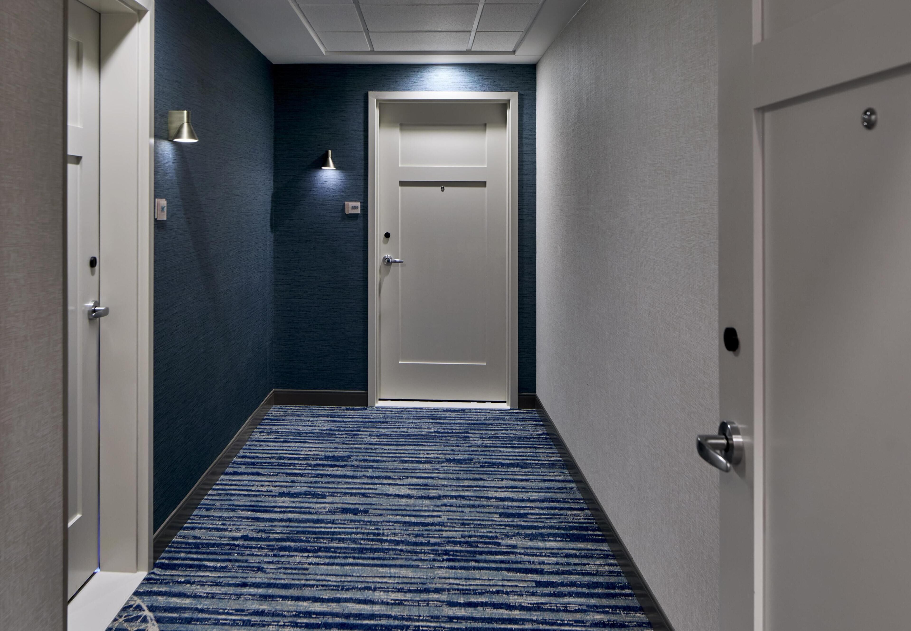 Un pasillo con alfombra azul, una puerta blanca y una lámpara fijada a la pared.