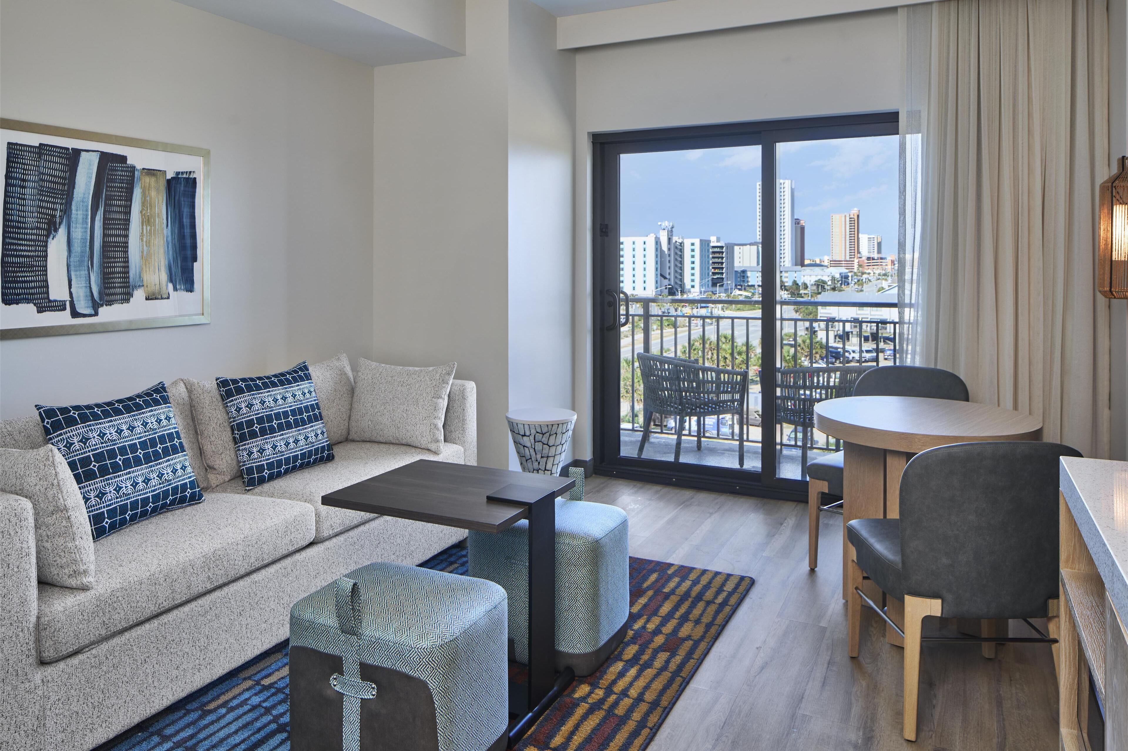 Una sala de estar moderna con sofá, reposapiés, mesa de centro y un comedor con mesa y sillas. Un amplio ventanal ofrece vistas de la ciudad.
