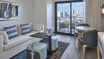 Un salon moderne comprenant un canapé, un pouf, une table basse et un coin repas avec une table et des chaises. Une grande fenêtre offre une vue sur le paysage urbain.