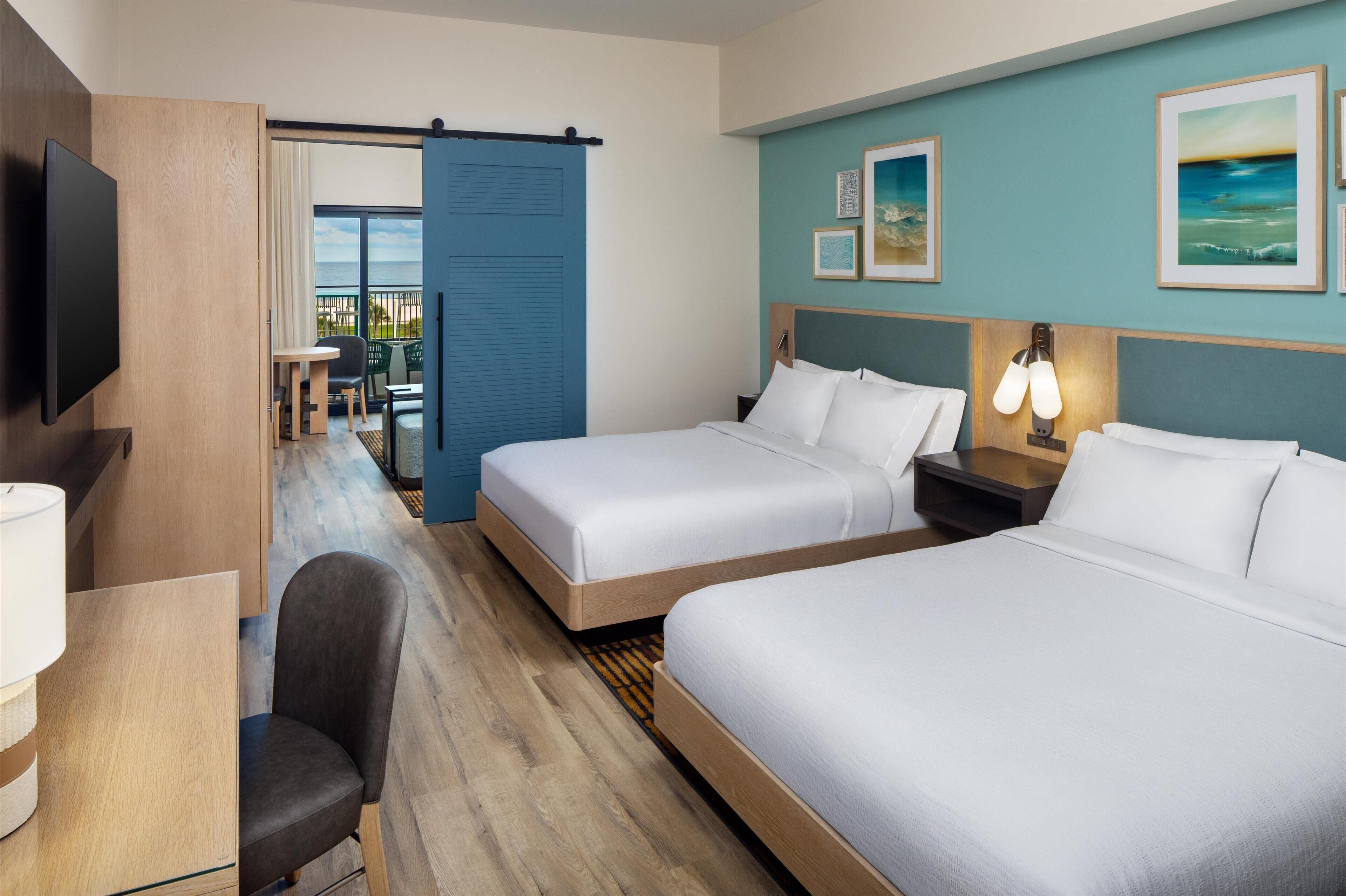 A hotel room with two beds, a desk with a chair, a TV, and a balcony with a view.