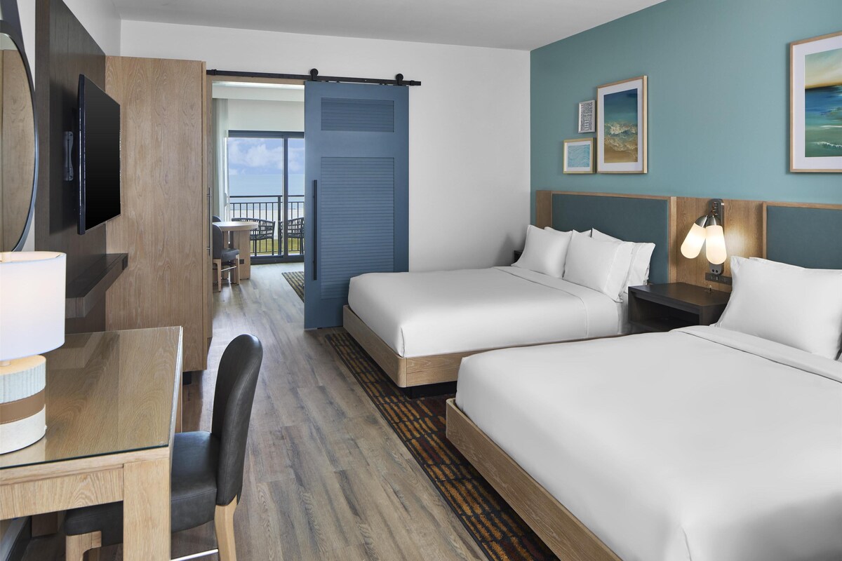 Ein Hotelzimmer mit zwei Betten, einem Schreibtisch, einem Stuhl, einem Fernseher und einem Balkon mit Blick.