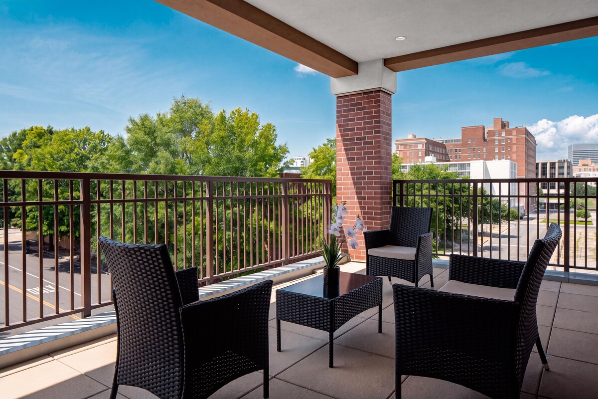 A balcony with wicker furniture and a potted plant, overlooking a street and buildings.
