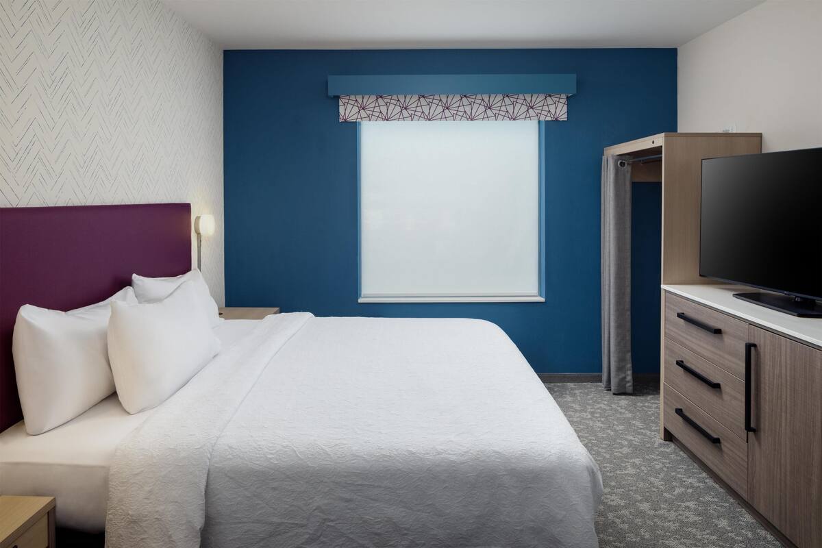 Una habitación de hotel con una cama, televisor, armario y ventana con cortina estampada.