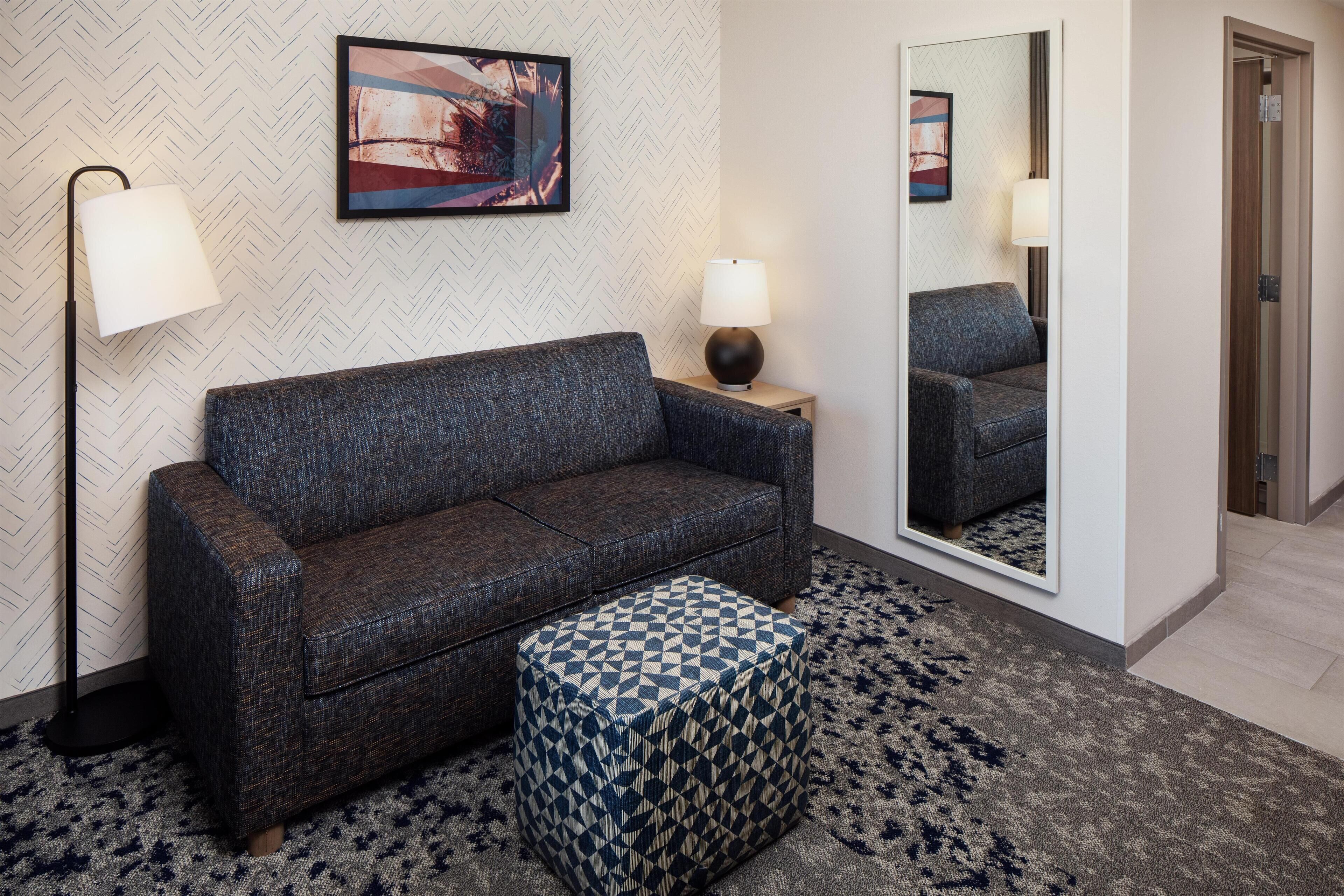Una habitación de hotel moderna con un sofá gris, un reposapiés estampado, una lámpara de pie y un espejo que refleja la habitación.