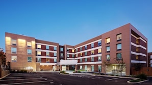 Un hotel de varios pisos con diseño moderno, que cuenta con una entrada central con galería techada, un estacionamiento y un asta de bandera.