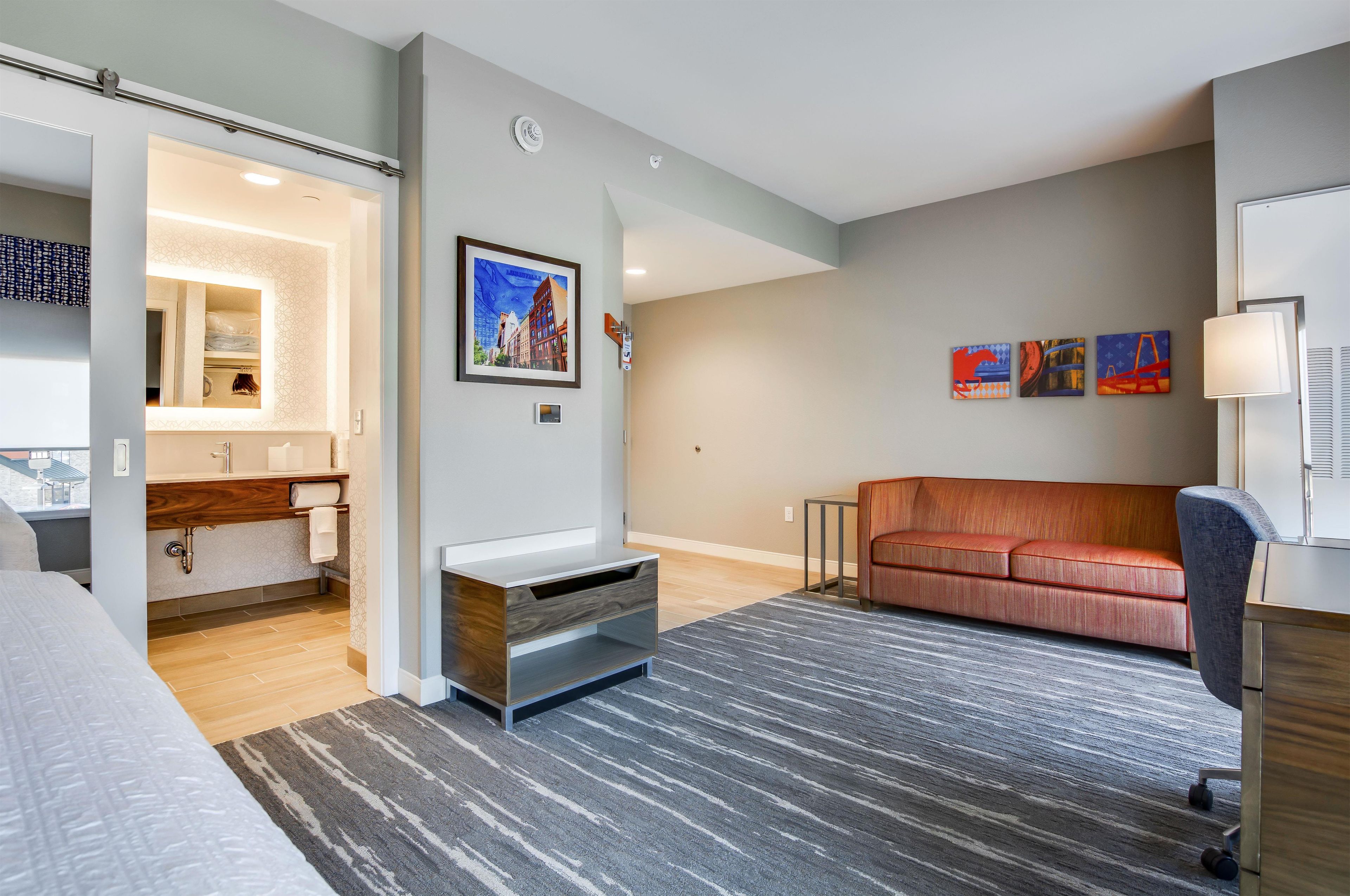 A modern hotel room with a bed, a sofa, a desk, a chair, a bathroom, and artwork on the walls.