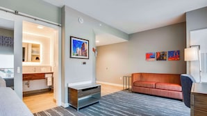 A modern hotel room with a bed, a sofa, a desk, and a bathroom with a vanity and mirror. - Hampton Inn & Suites By Hilton Louisville East Oxmoor (Louisville)