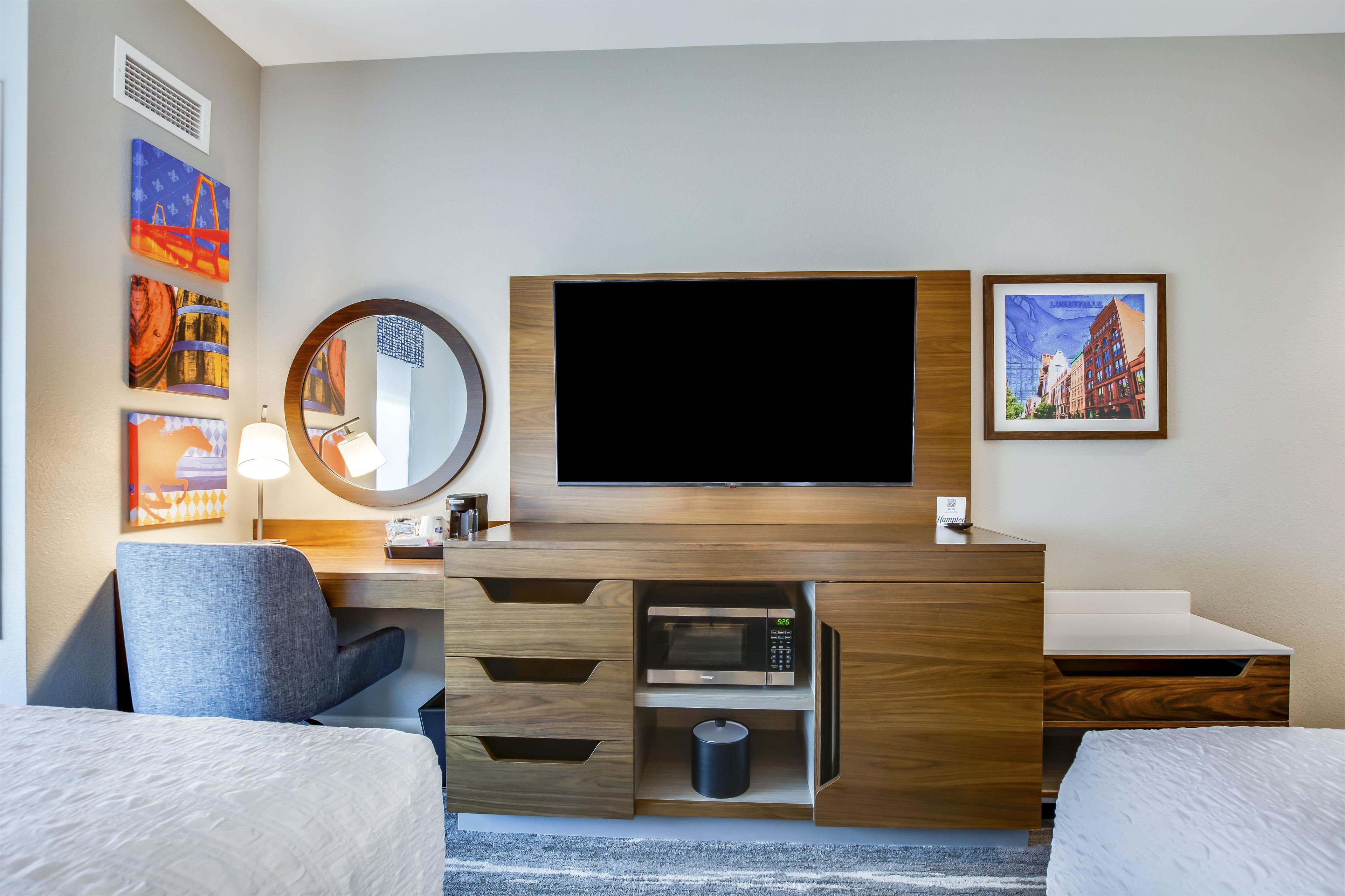 A hotel room with a flat-screen TV, a desk with a lamp, a mirror, and artwork on the walls.