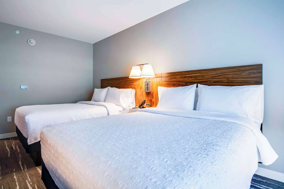 Habitación de hotel con dos camas, cabecera de madera, lámpara de pared y suelo cubierto de alfombra.