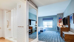 A modern hotel room with a sliding door leading to a bathroom, a desk with a chair, a bed, and a large window with a view of buildings. - Hampton Inn & Suites By Hilton Louisville East Oxmoor (Louisville)