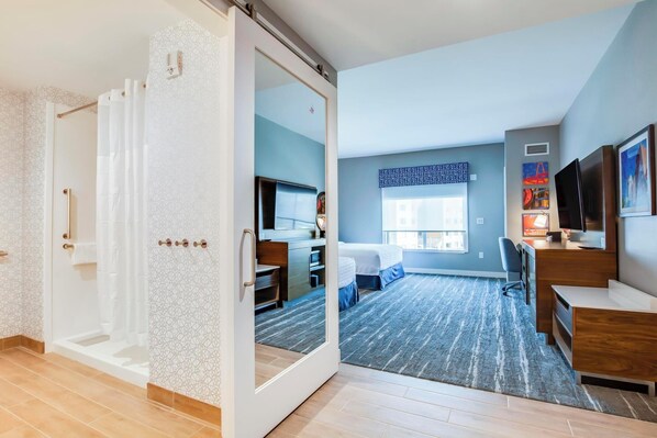 Une chambre d’hôtel moderne dotée d’une porte coulissante donnant accès à la salle de bain, d’un bureau avec une chaise, d’un lit et d’une grande fenêtre offrant une vue sur les bâtiments.