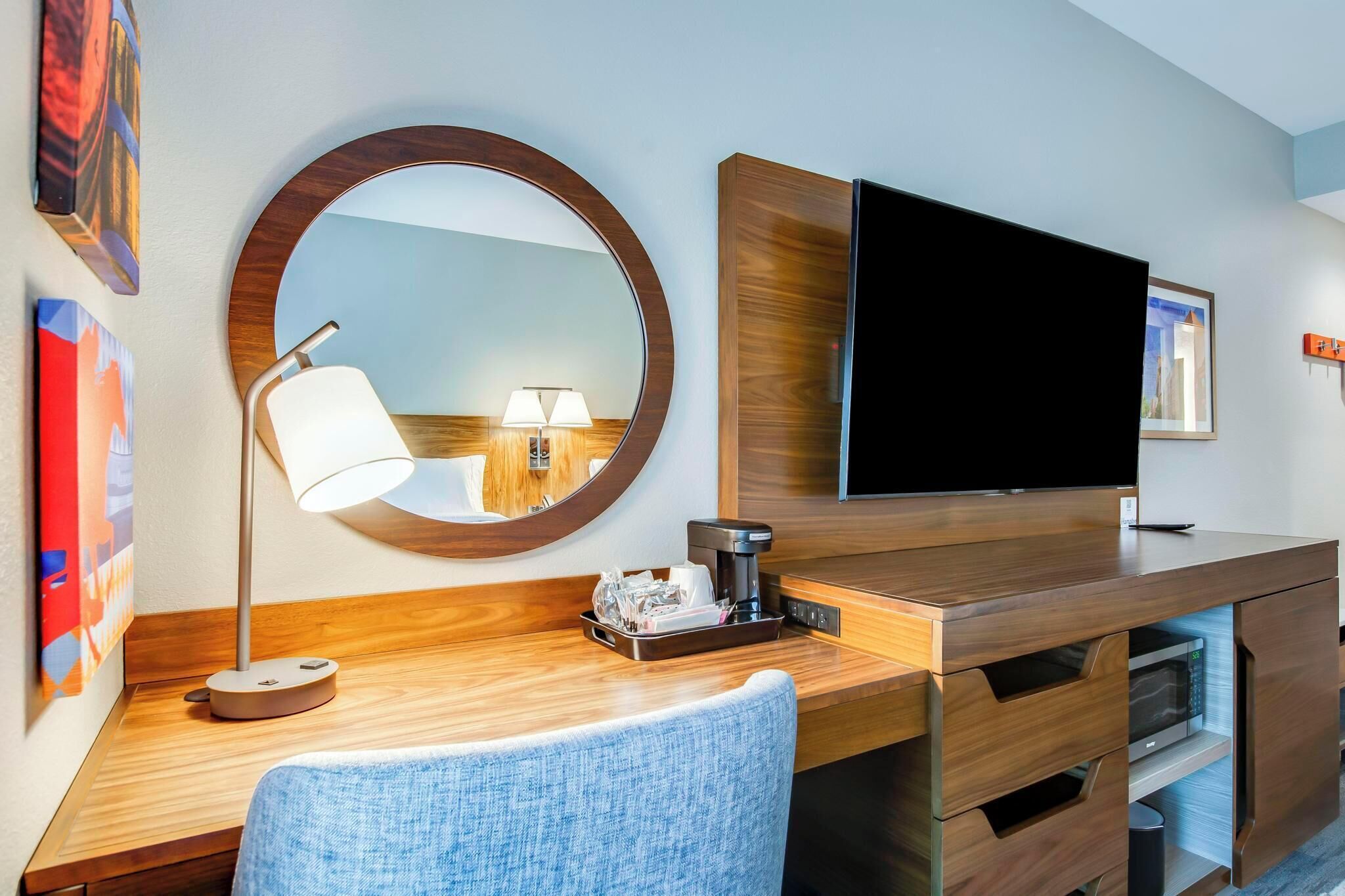 A hotel room with a wooden desk, a round mirror, a flat-screen TV, a lamp, and a bed.