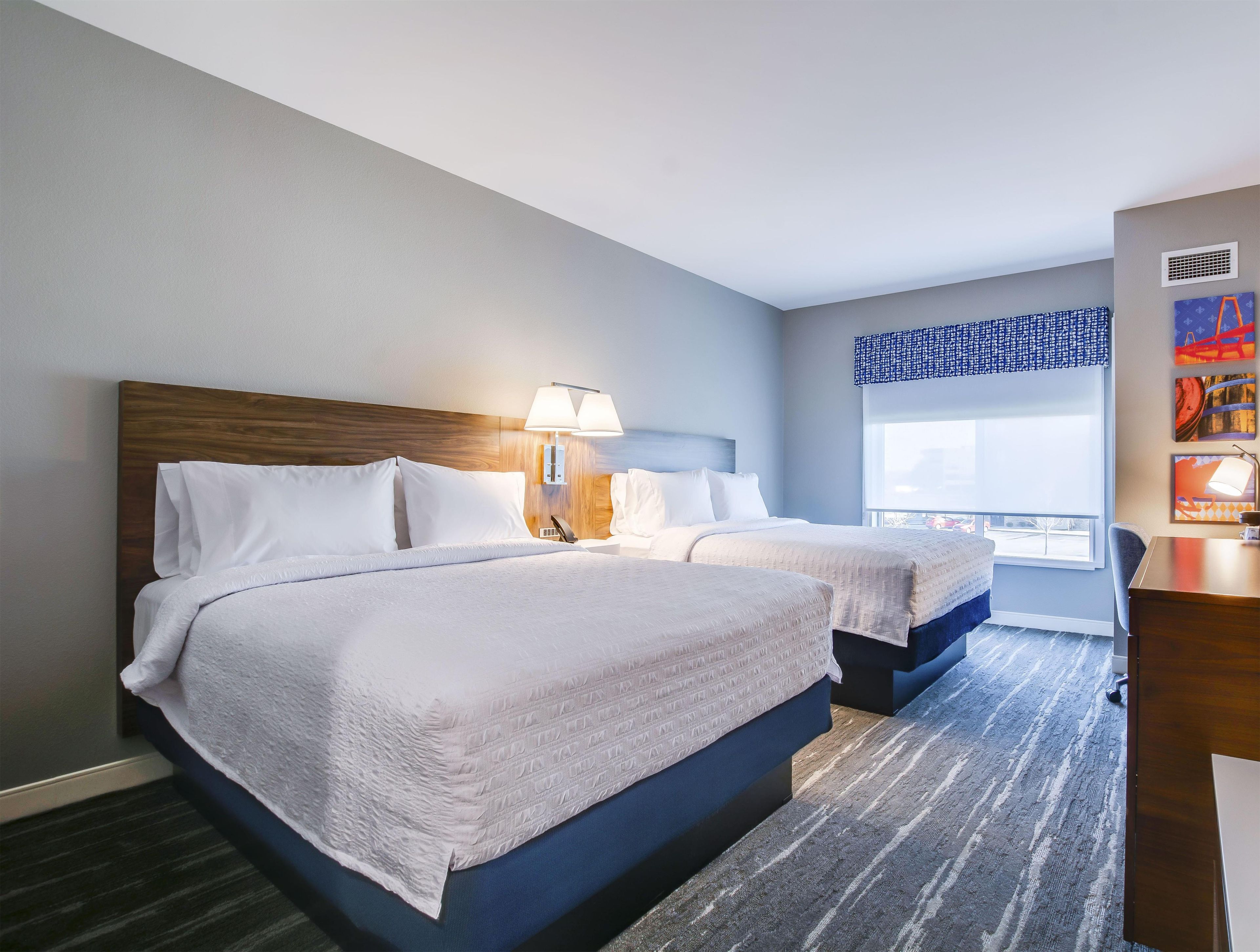 A hotel room with two beds, a desk, a window with a view, and a painting on the wall.