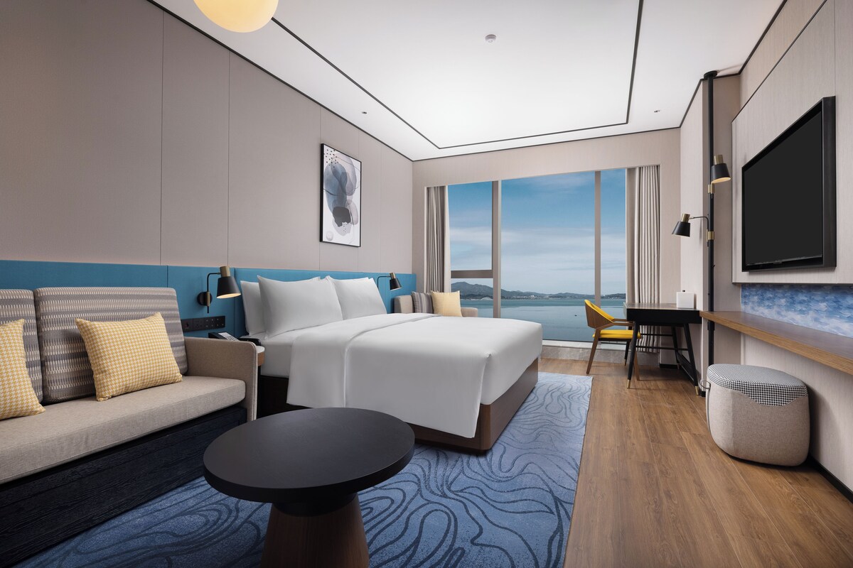 A modern hotel room with a bed, sofa, desk, and a large window offering a view.