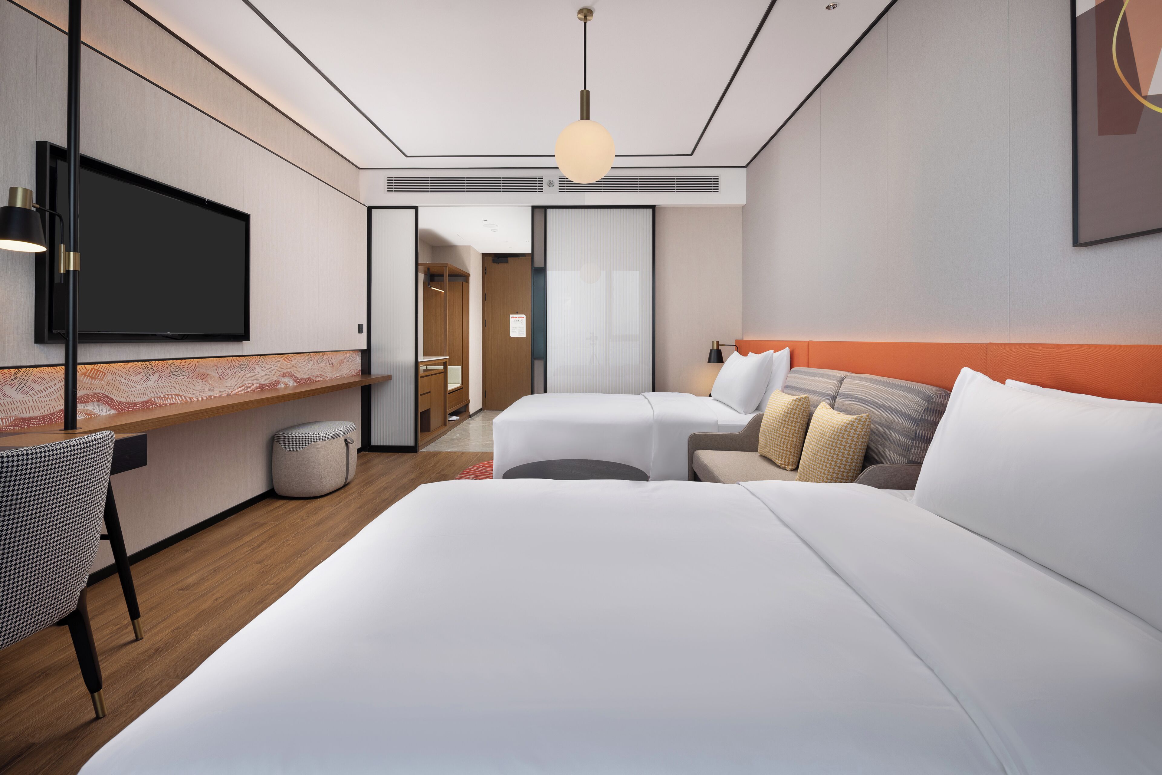 Une chambre d’hôtel moderne dotée d’un grand lit, d’un bureau avec un téléviseur à écran plat, d’un coin salon avec un canapé et un pouf, et d’un couloir menant à la salle de bain.